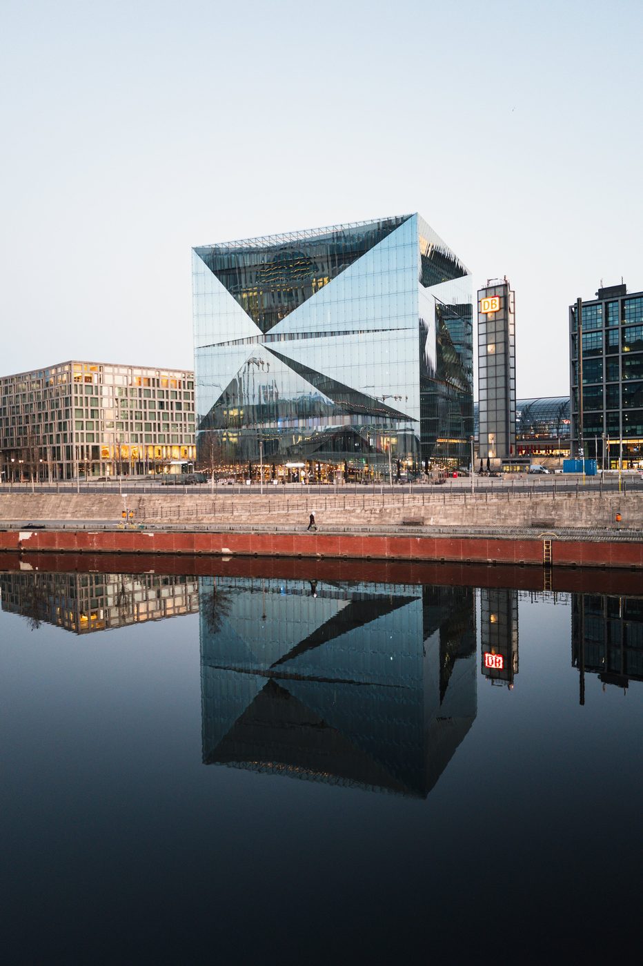 Cubic glass building and its reflection in the canal, a passerby walking along the quay, Berlin