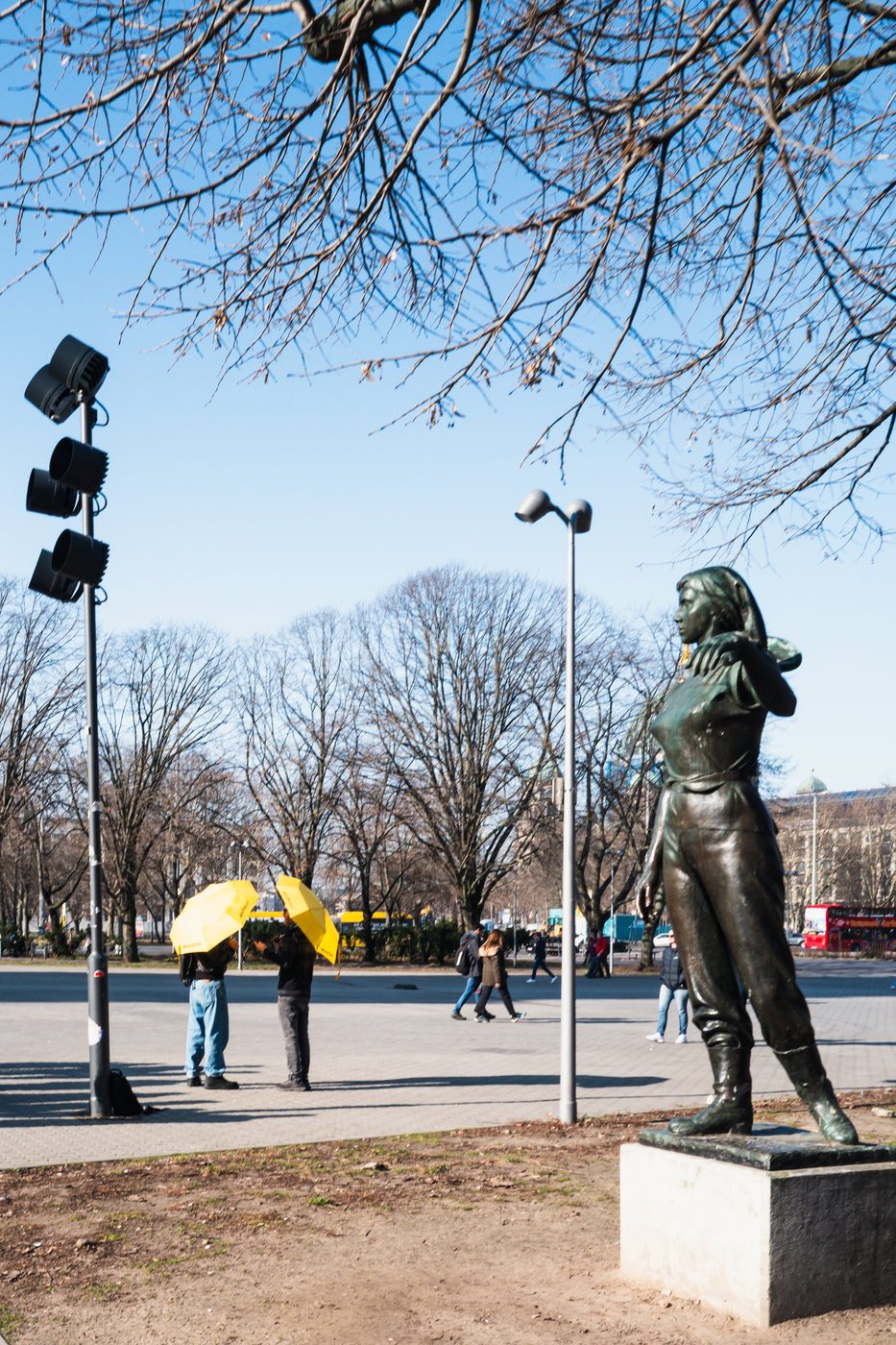 Bronze statue facing two passersby beneath yellow umbrellas, Berlin