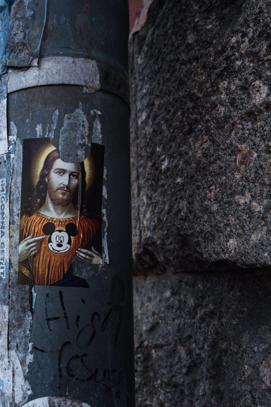 Altered sticker on a pole showing a religious figure's face masked by Mickey Mouse, Berlin