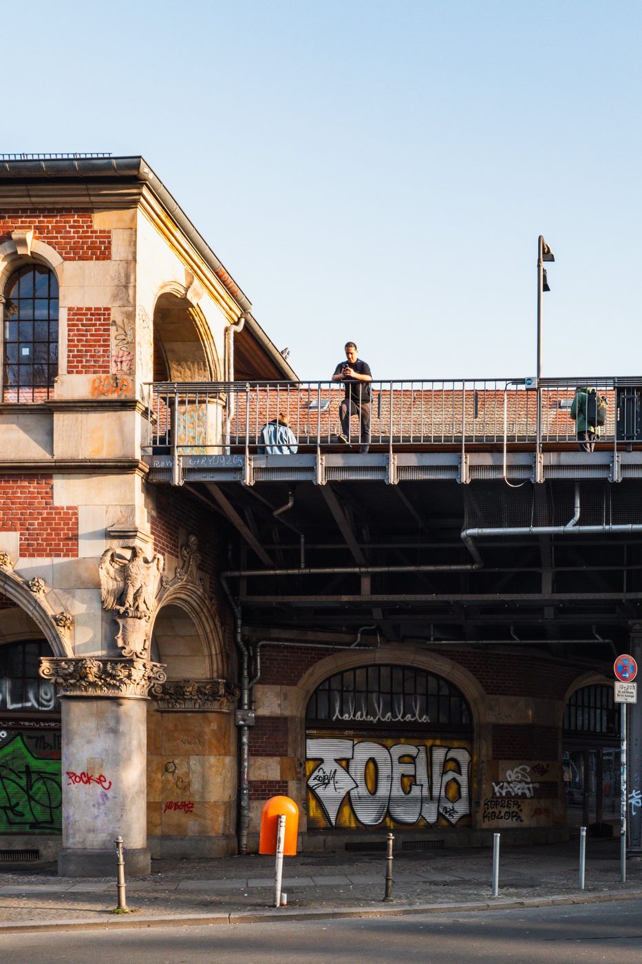 Brick railway bridge looming over a tagged archway, a passerby leaning against it, Berlin
