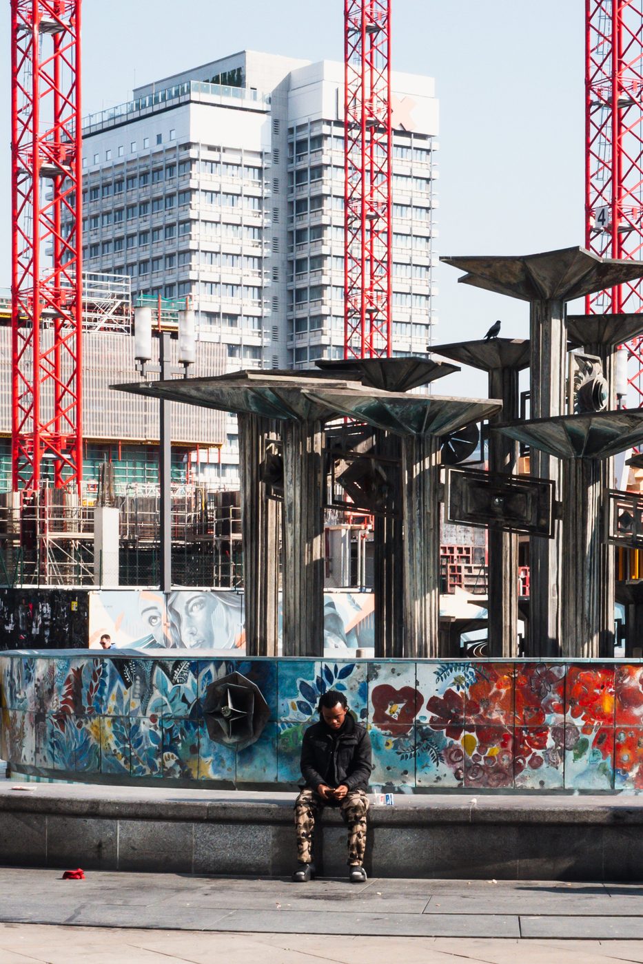 Man sitting on a mosaic fountain facing red construction cranes, Berlin