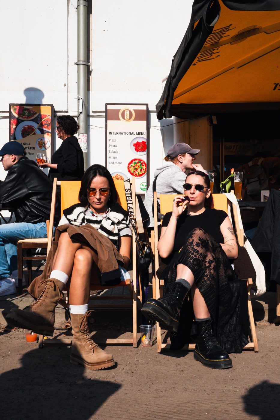 Two women in deck chairs at the market, wearing boots and sunglasses, Berlin