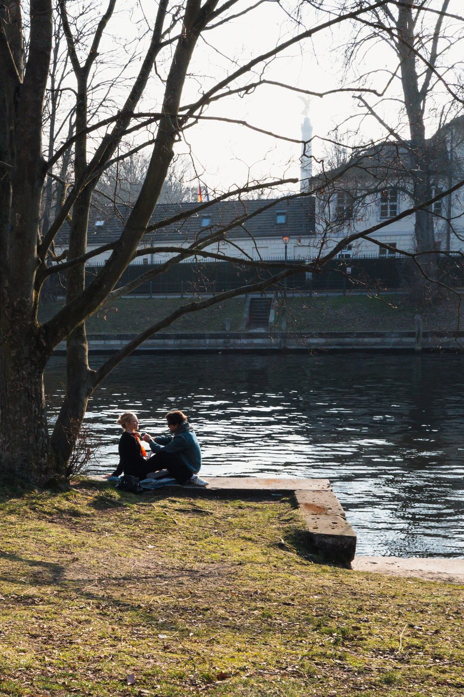 Couple sitting by the water beneath a willow, winged statue in the distance, Berlin