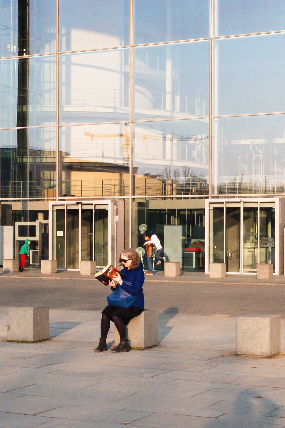 Woman reading a book seated on a concrete block in front of a glass facade, Berlin