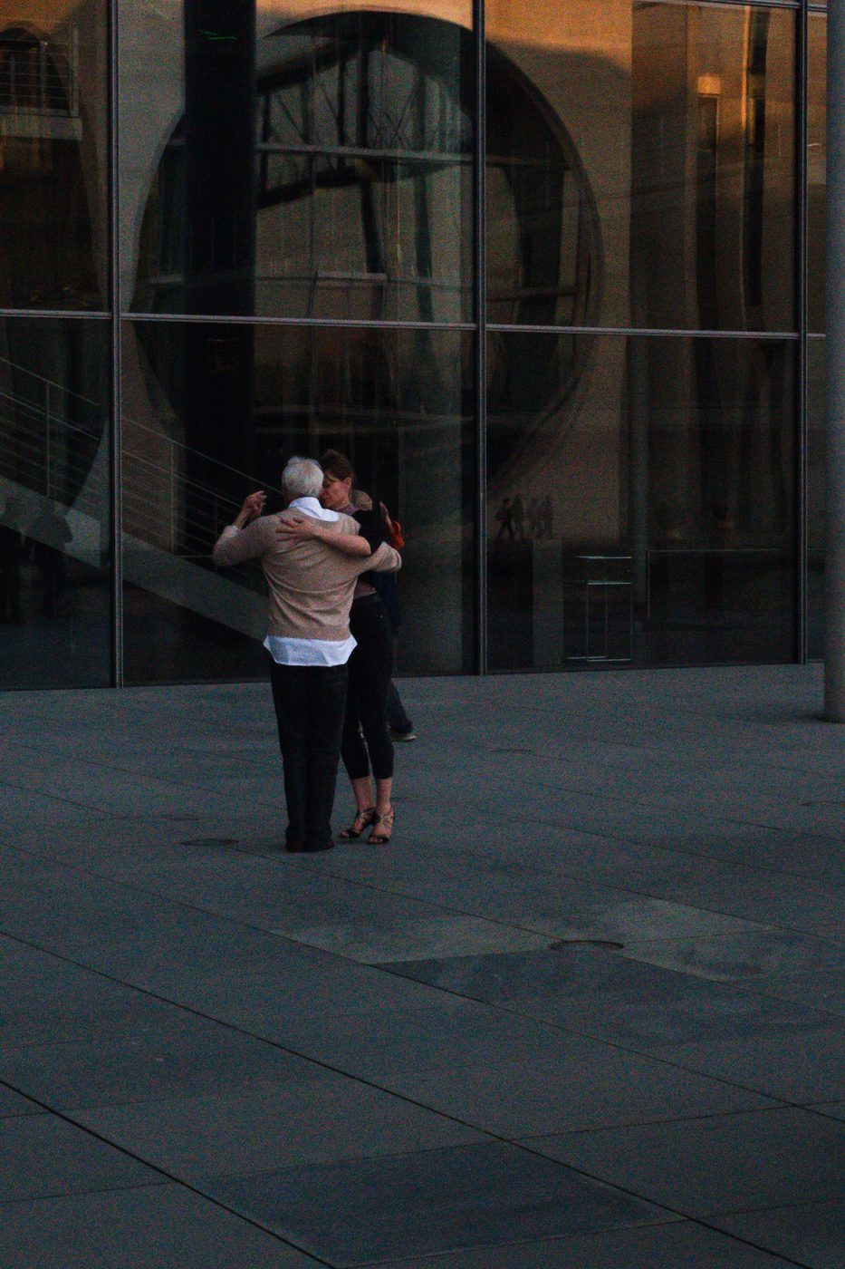 Elderly couple dancing embraced in front of a glass facade at dusk, Berlin