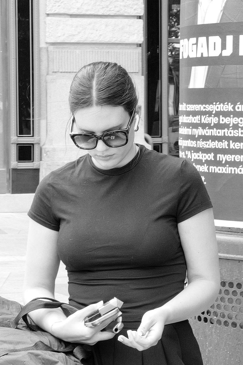 A woman checking her phone, sunglasses and a lottery poster, Budapest