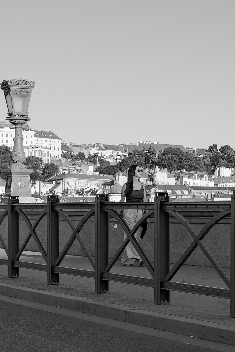 A woman in sunglasses walking along a bridge near an old lamppost, Budapest