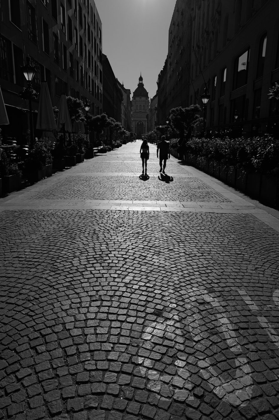Two people crossing a cobbled street backlit toward a basilica, Budapest