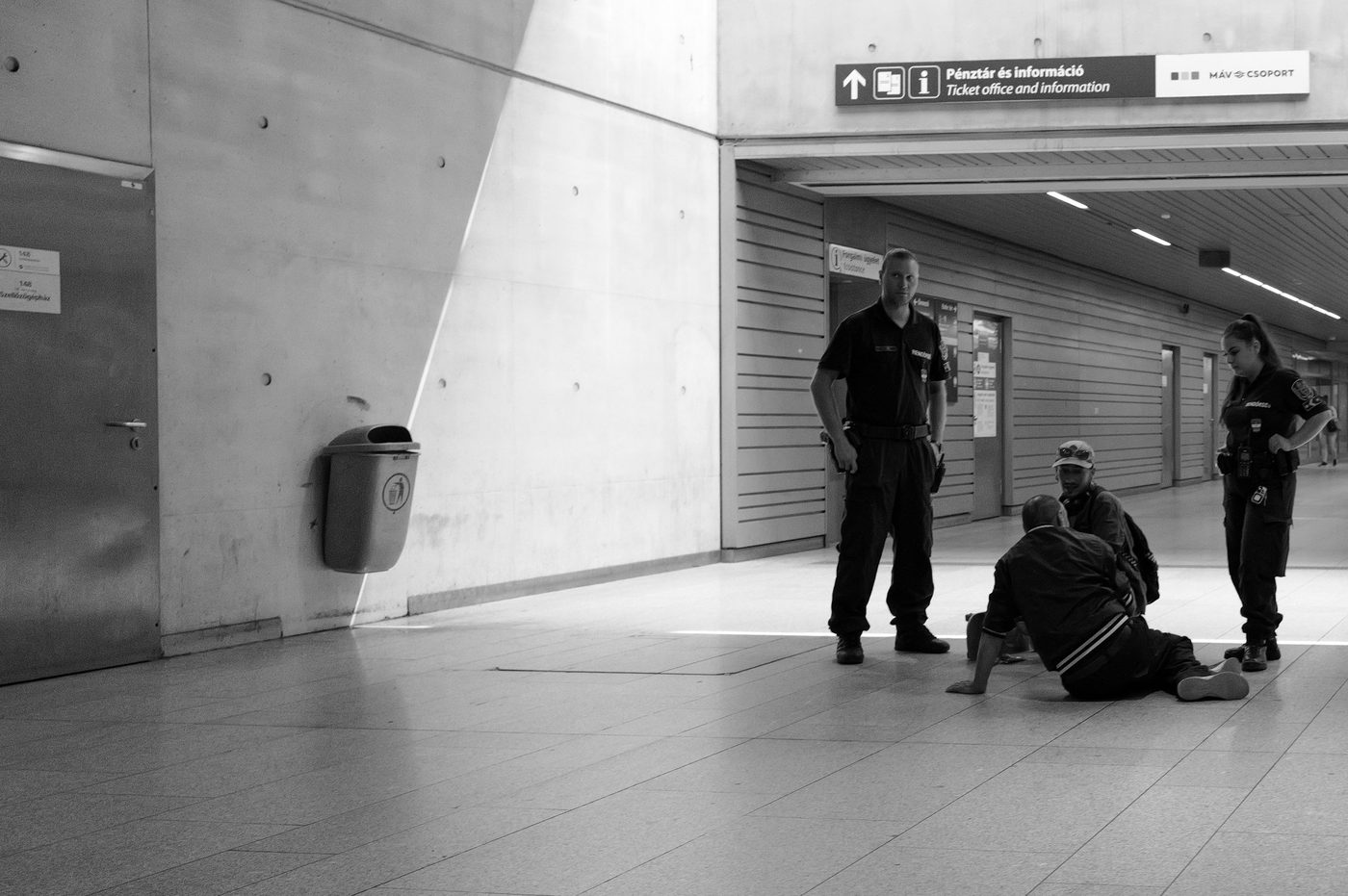Security officers attending to a man seated on the ground in a station hall, Budapest