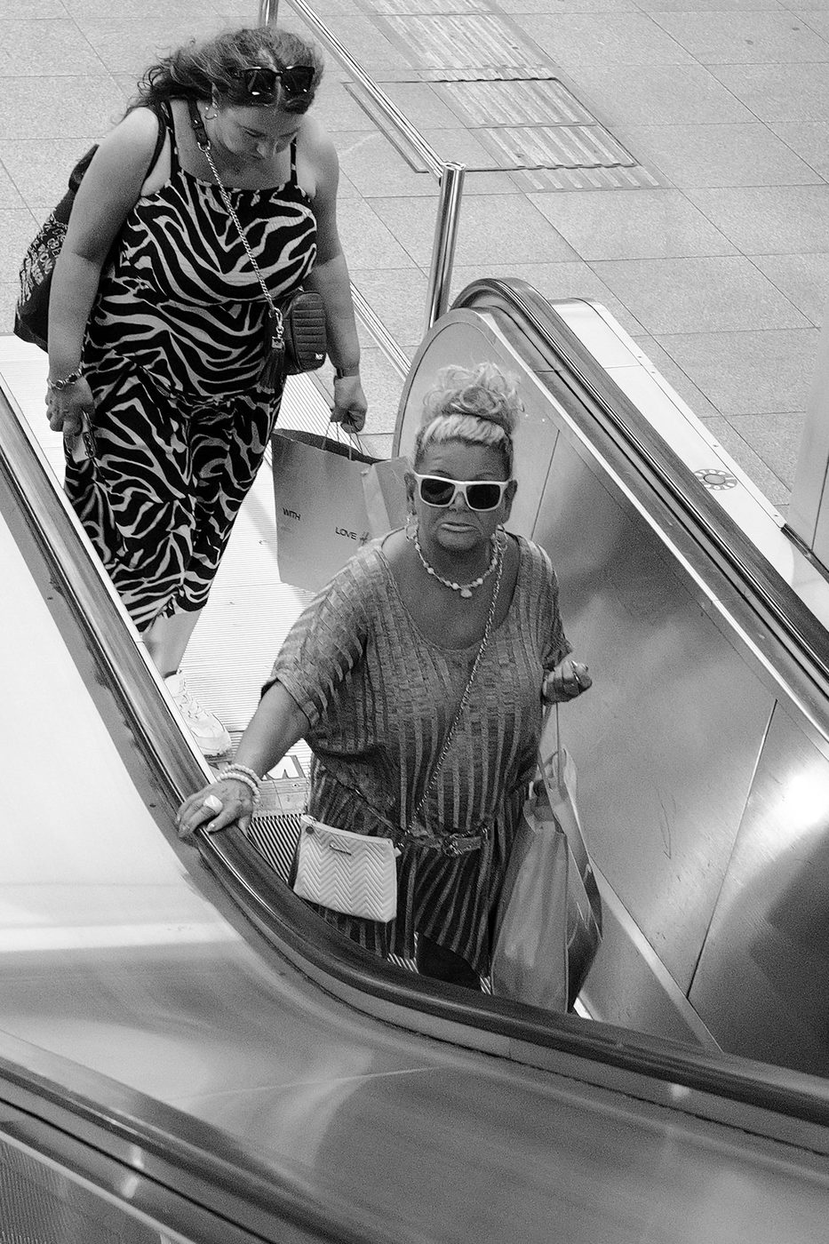 Two women in printed outfits going down an escalator, Budapest