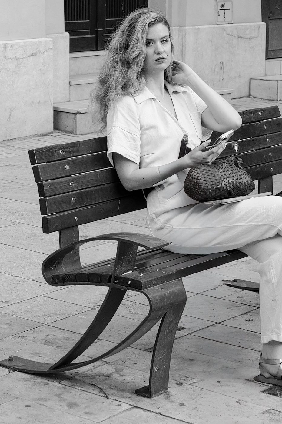 A woman with wavy hair sitting on a bench, phone in hand, Budapest