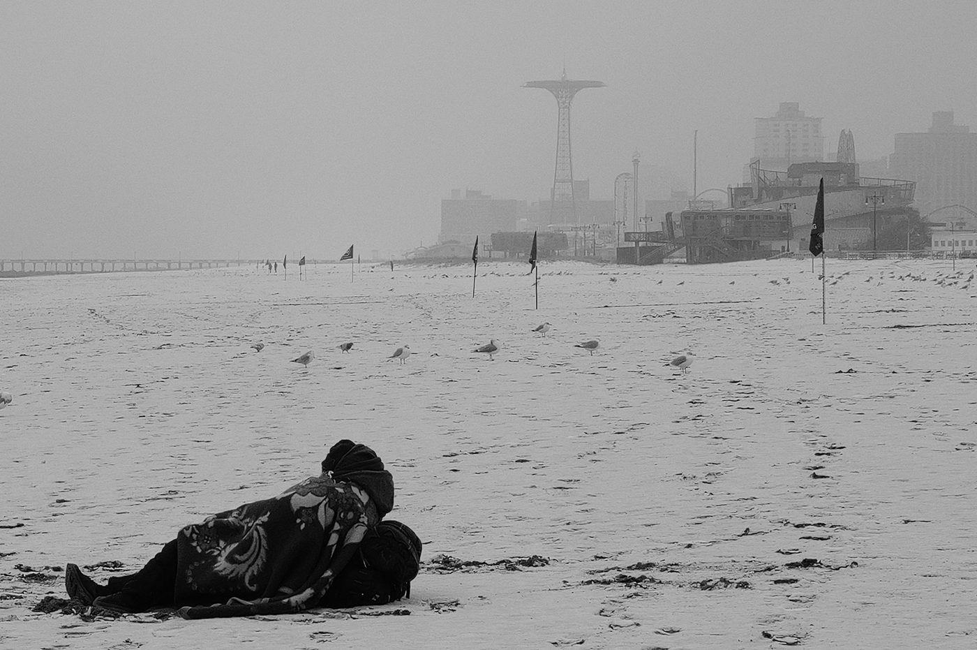 Person wrapped in a blanket on the snow-covered beach, Coney Island parachute tower in the background