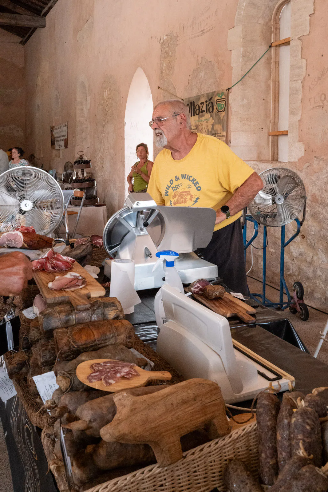 Un charcutier en tee-shirt jaune derrière son étal, sous une halle de pierre, Bonifacio, Corse