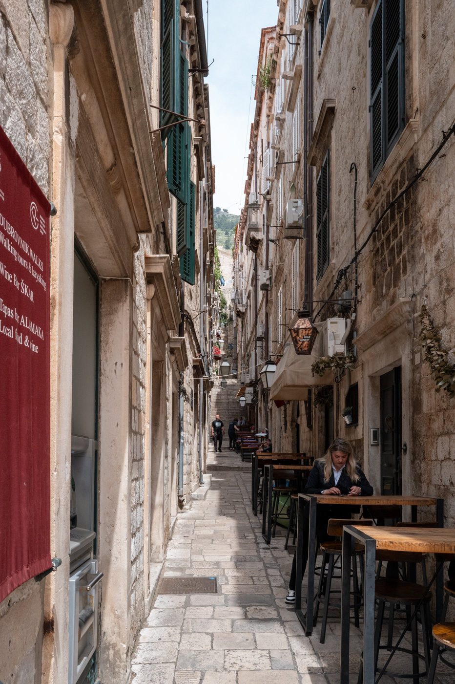Woman sitting at a high table in a sloping alley, Dubrovnik