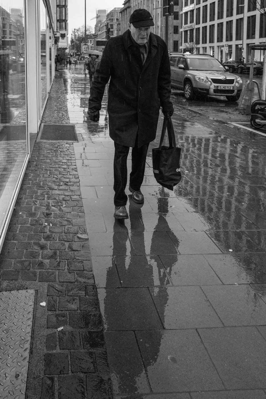 An elderly man walking in the rain with a bag, reflections on the ground, Düsseldorf