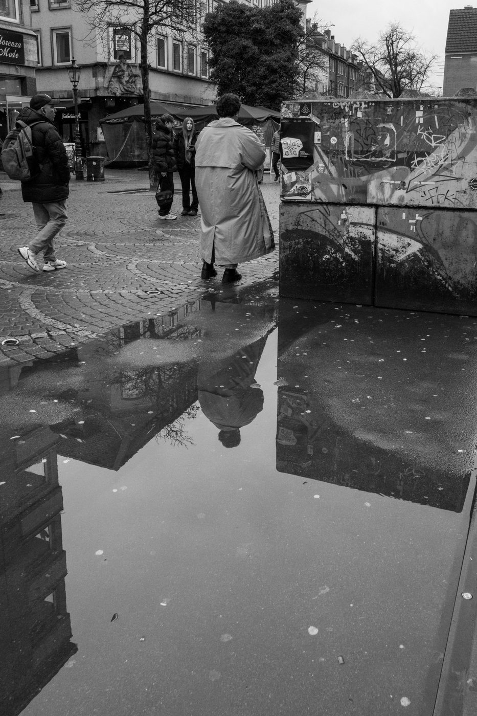 A woman in a trench coat reflected in a puddle, paved square, Düsseldorf