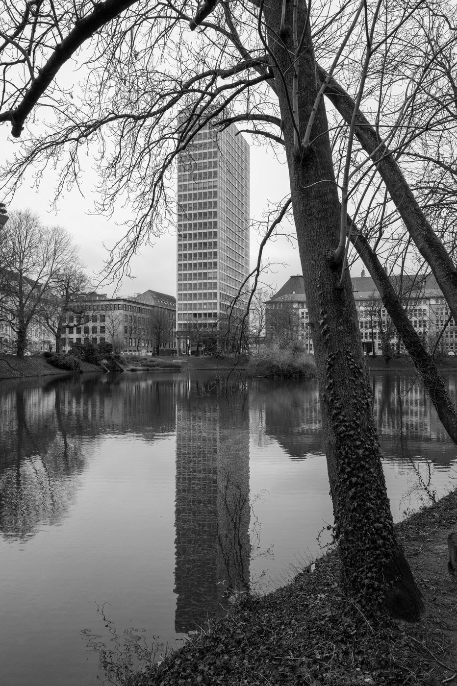A slender glass tower reflected in a body of water lined with trees, Düsseldorf