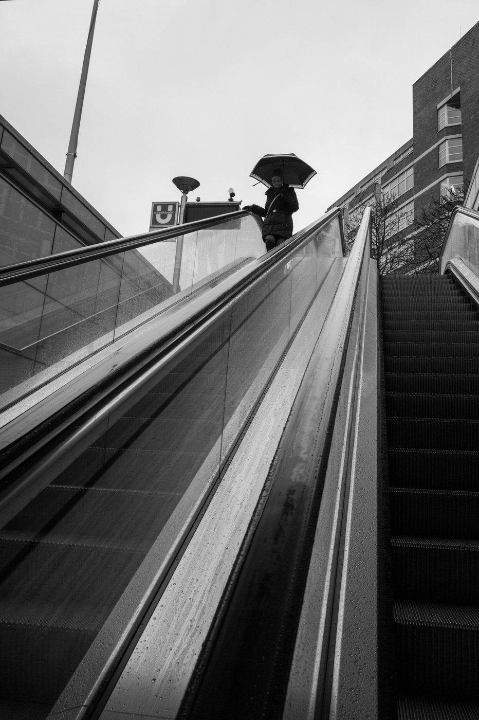 A woman under an umbrella at the top of an escalator, brick building, Düsseldorf