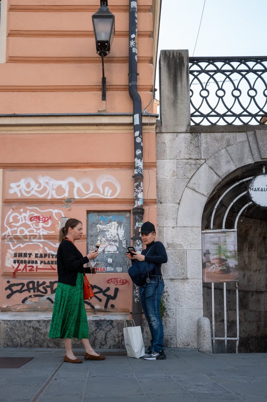 Two women checking their phones in front of a graffiti-covered wall, Ljubljana