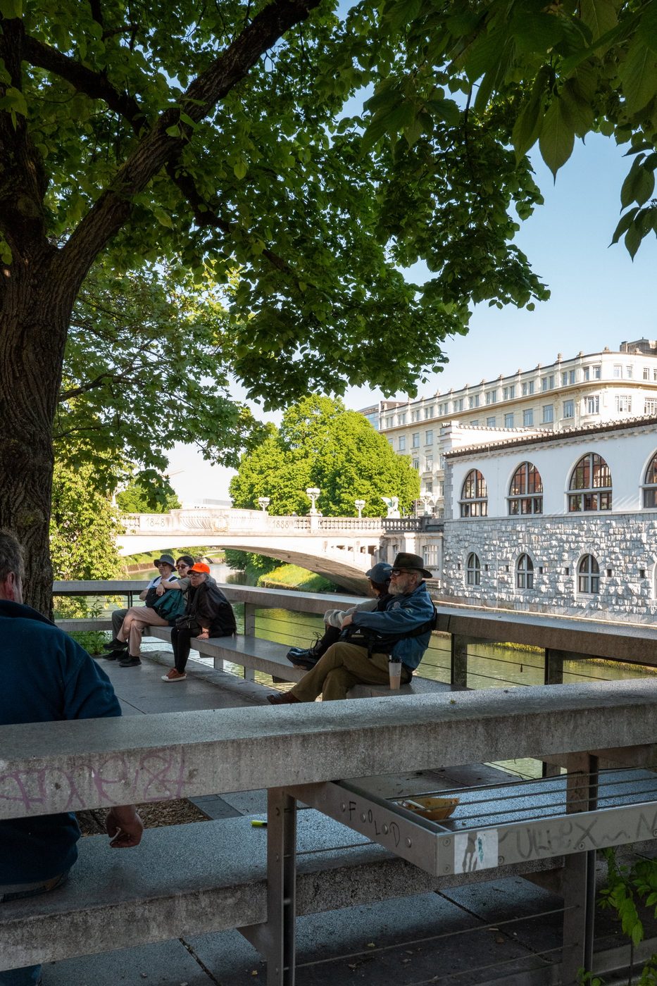 Tourists sitting under a tree facing the bridge and the arcades, Ljubljana