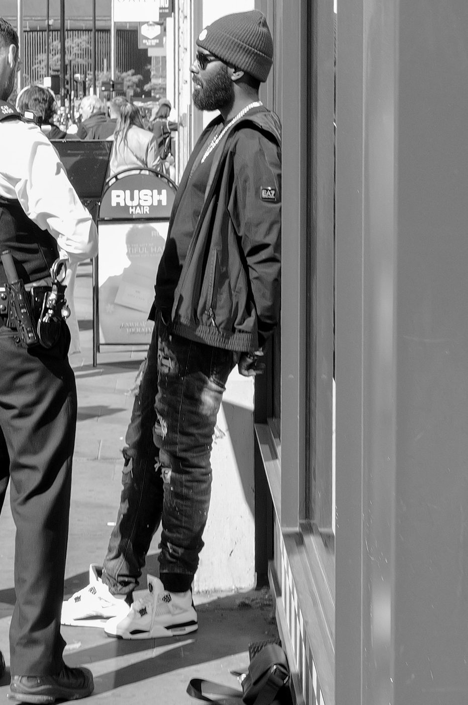A man in a beanie leaning against a shop window, police officer in the foreground, London