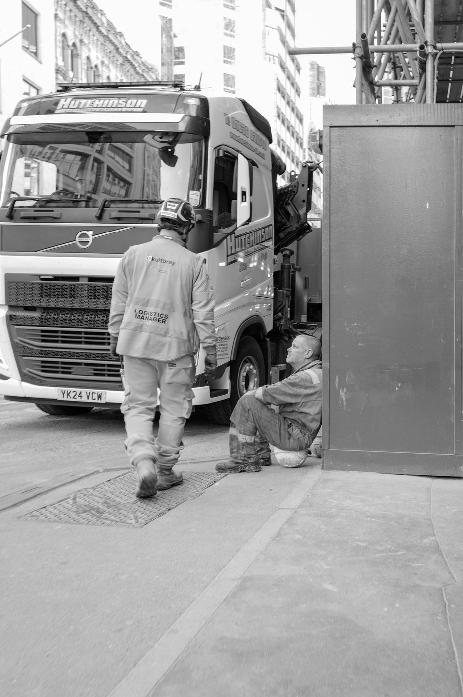 Worker in a logistics vest walking in front of a truck, colleague crouching near the site, London