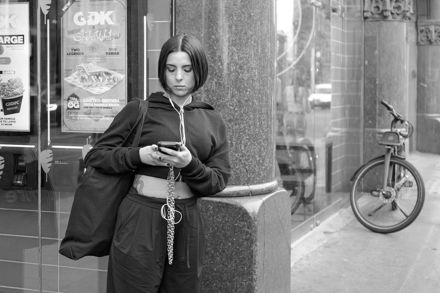 Young woman with a bob haircut, wired earphones, absorbed in her phone near a bicycle, London