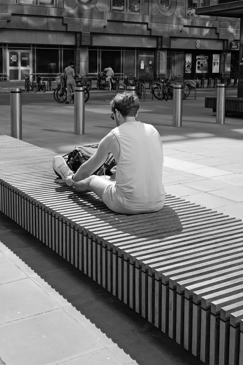 Man sitting cross-legged on a wooden-slat bench, busy modern square, London