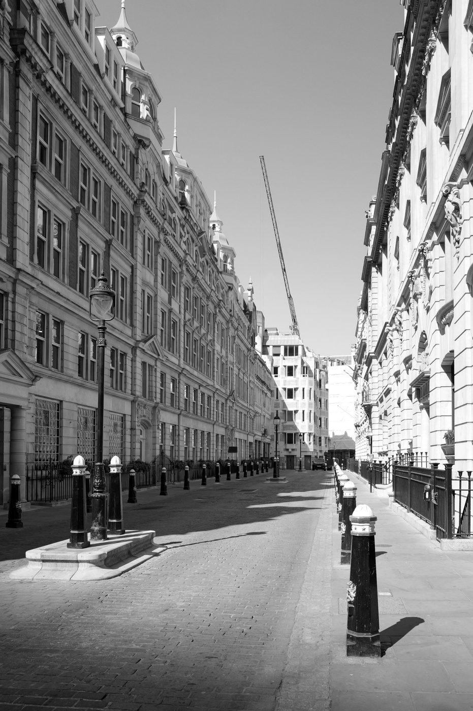 Deserted historic street lined with black bollards, ornate buildings and a crane in the distance, London