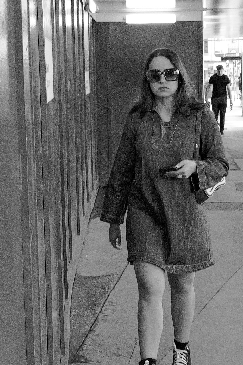 Young woman in a denim shirt dress crossing a covered passage, phone in hand, London