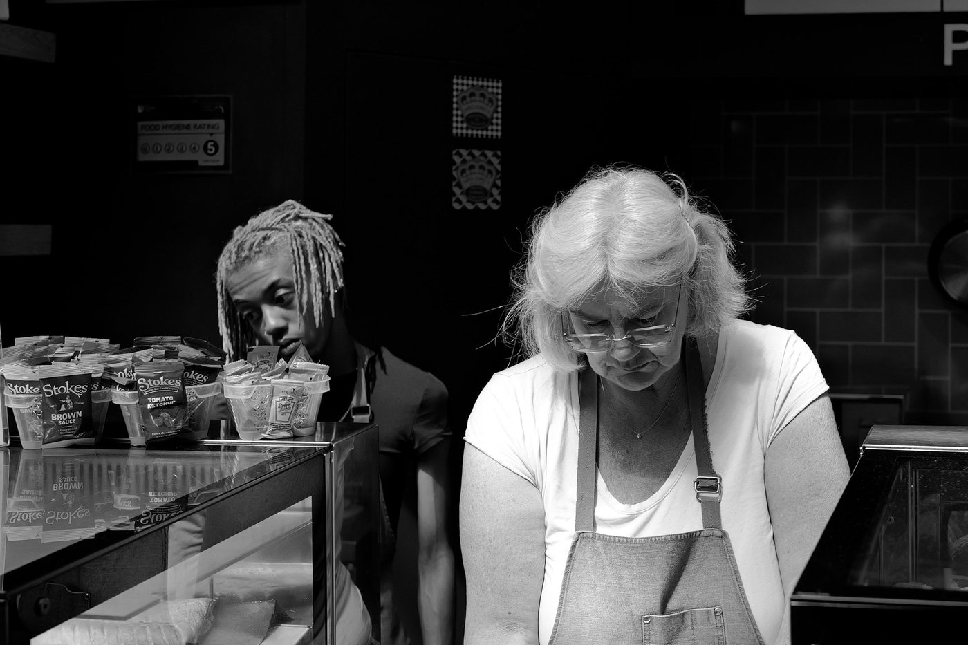 Saleswoman in an apron leaning over her counter, young man with braids in the background, London