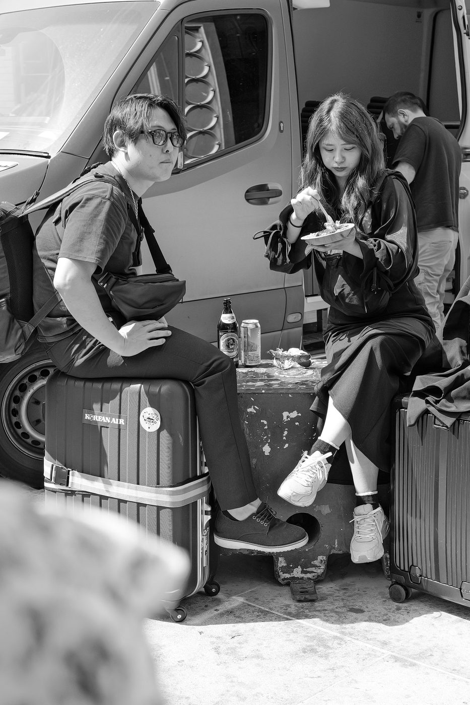 Two travellers sitting on their suitcases sharing street food near a van, London