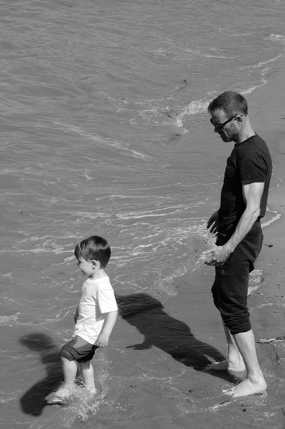 A father watches his young son paddle barefoot in the waves, London