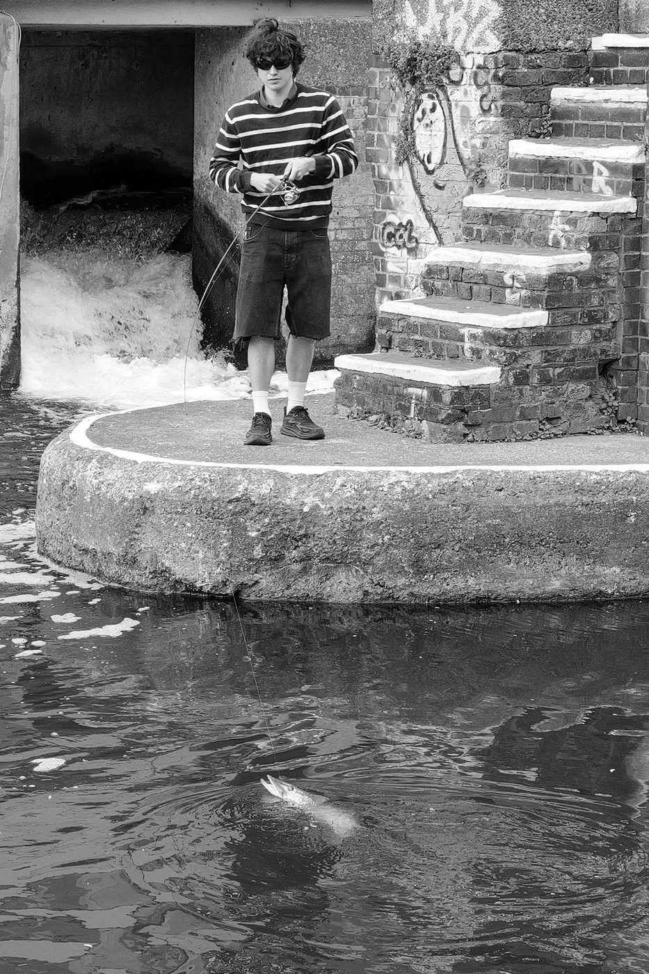 Young fisherman in shorts reeling in a fish from the canal's concrete bank, London