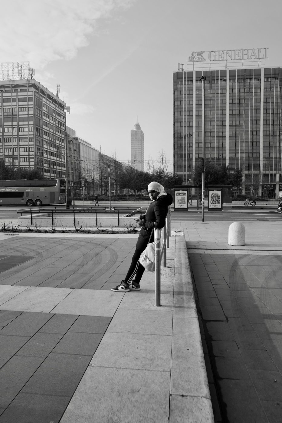 Young woman in a headscarf leaning against a railing, phone in hand, modern towers in the background, Milan