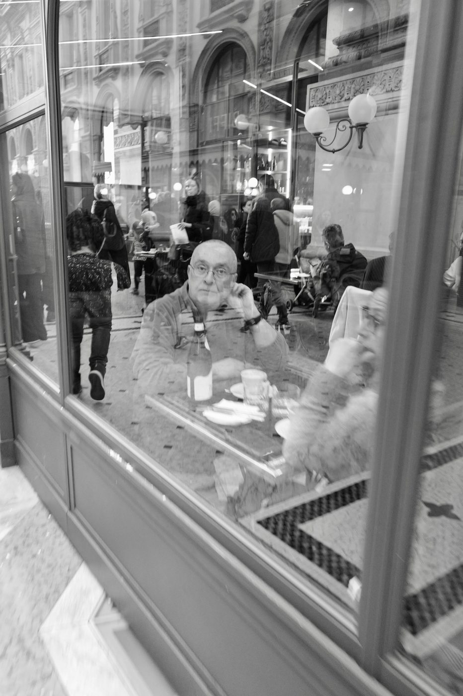 Reflection in a shop window of a man alone at a cafe table, a bottle of wine in front of him, Milan