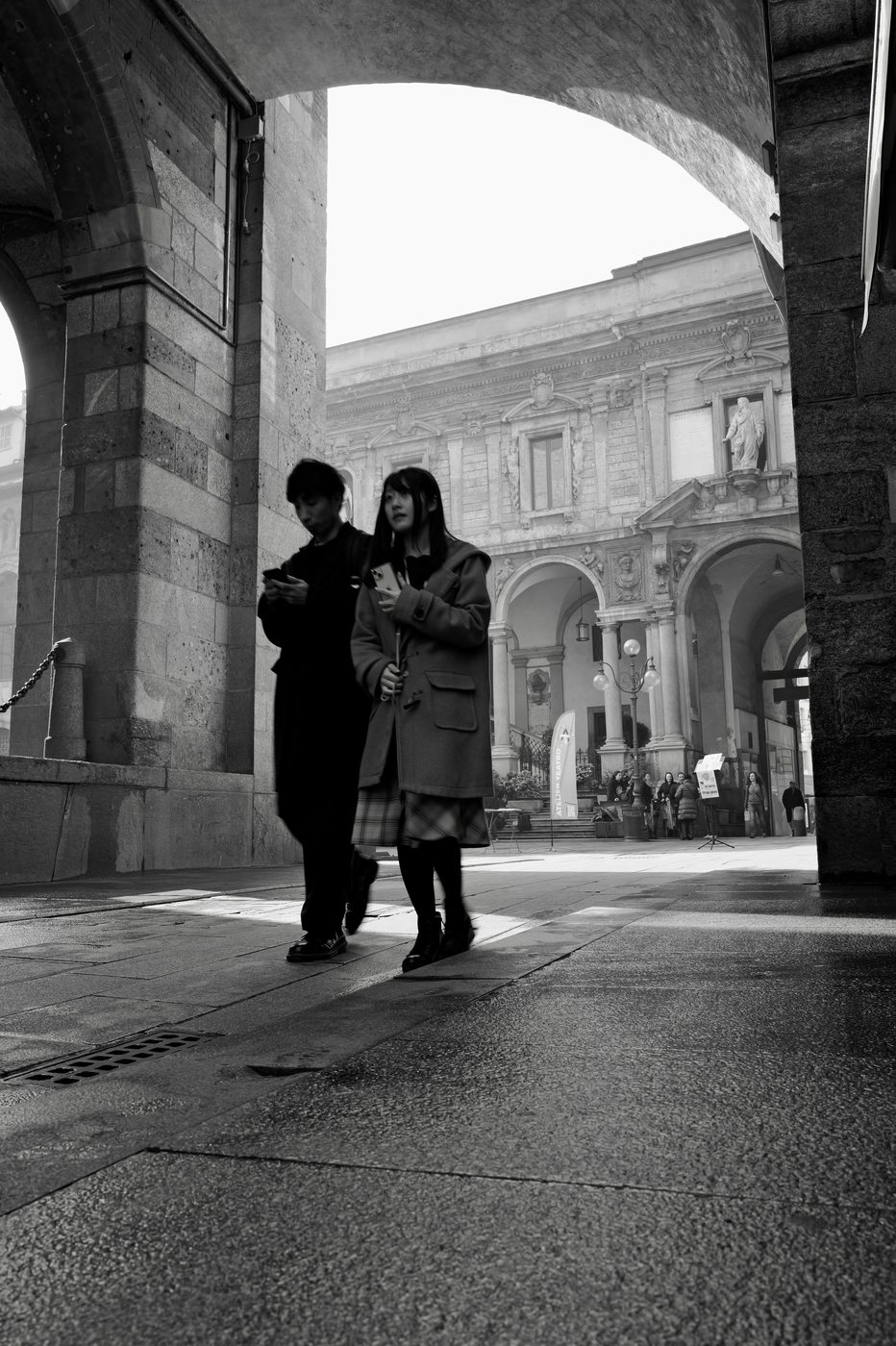 Silhouettes of a couple walking under an arcade, absorbed in their phones, Milan
