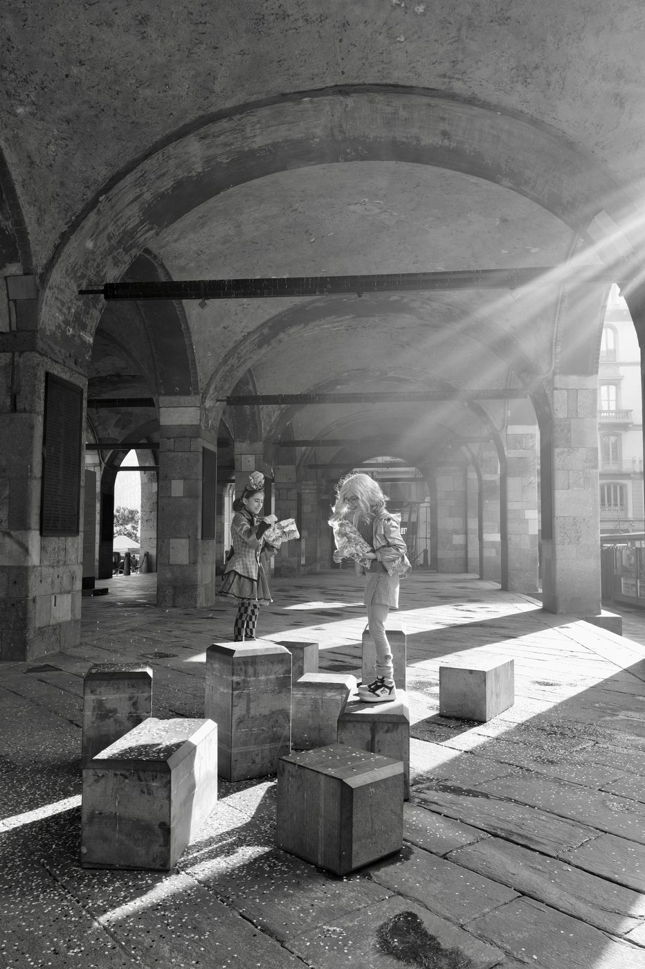 Two children perched on stone blocks, rays of light crossing an arcade, Milan
