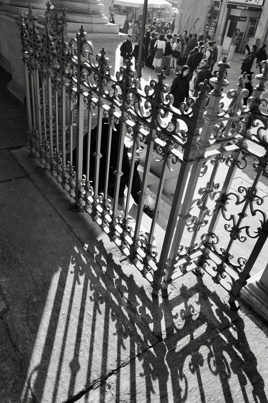 Wrought-iron gate casting its shadows on the pavement, blurred crowd in the background, Milan