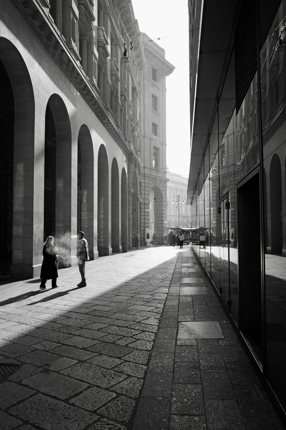Two passers-by talking under an arcade, breath visible in the cold air, Milan