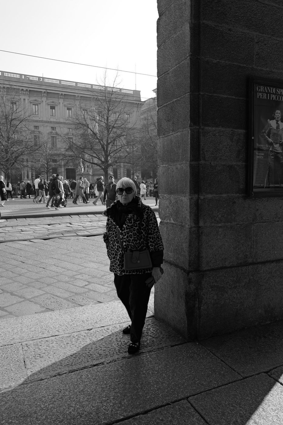 Elderly woman in a leopard-print coat passing a poster for a La Scala performance, Milan