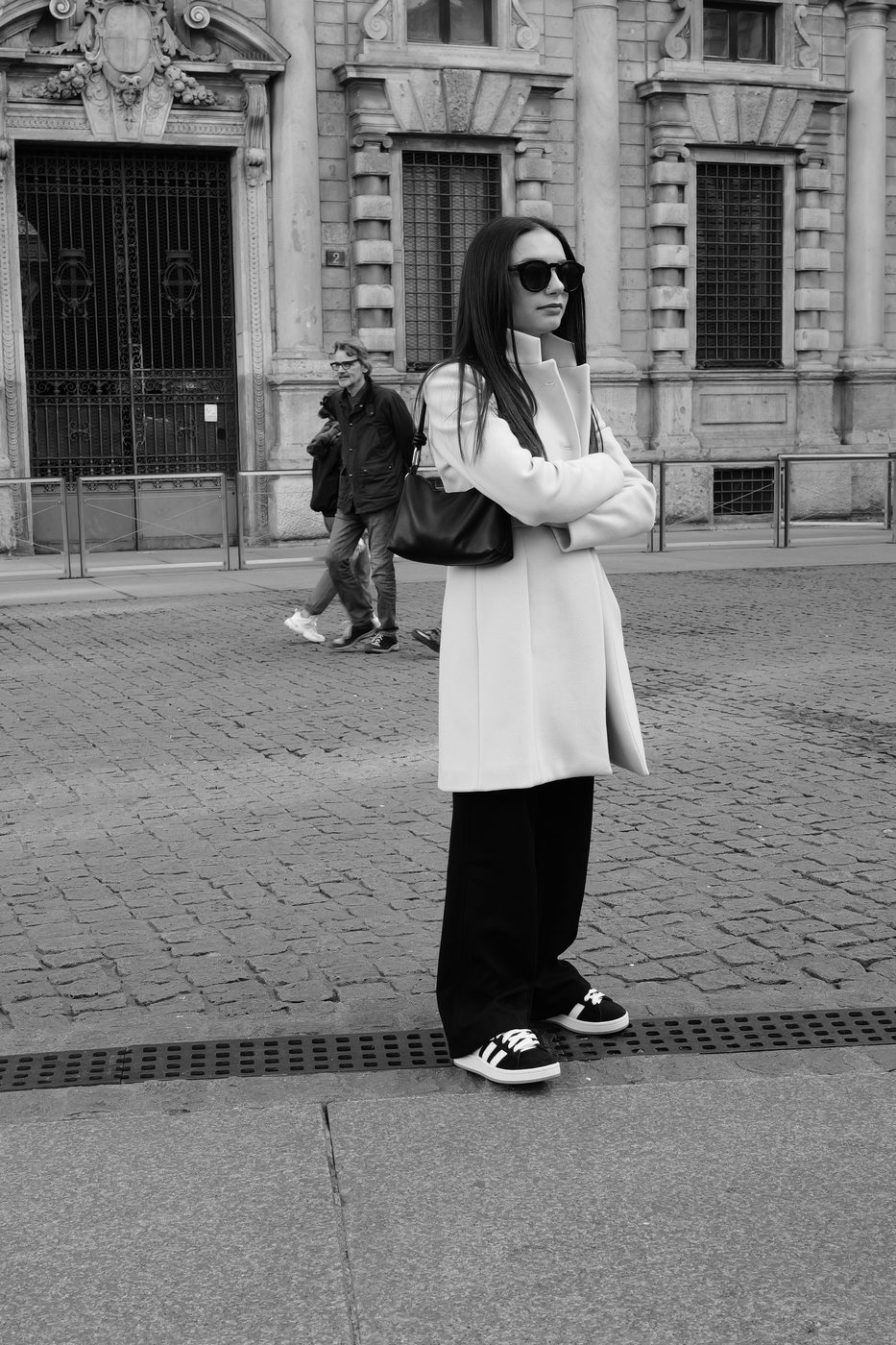 Woman in a white coat and dark sunglasses, arms crossed on a cobbled square, Milan