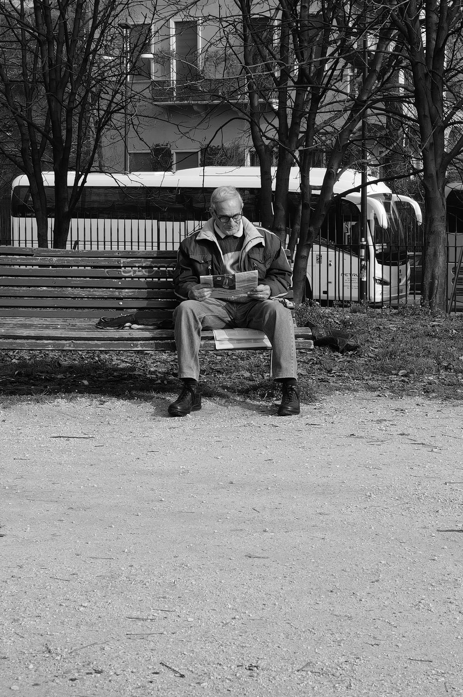 Elderly man reading his newspaper on a public bench, in front of a parked coach, Milan