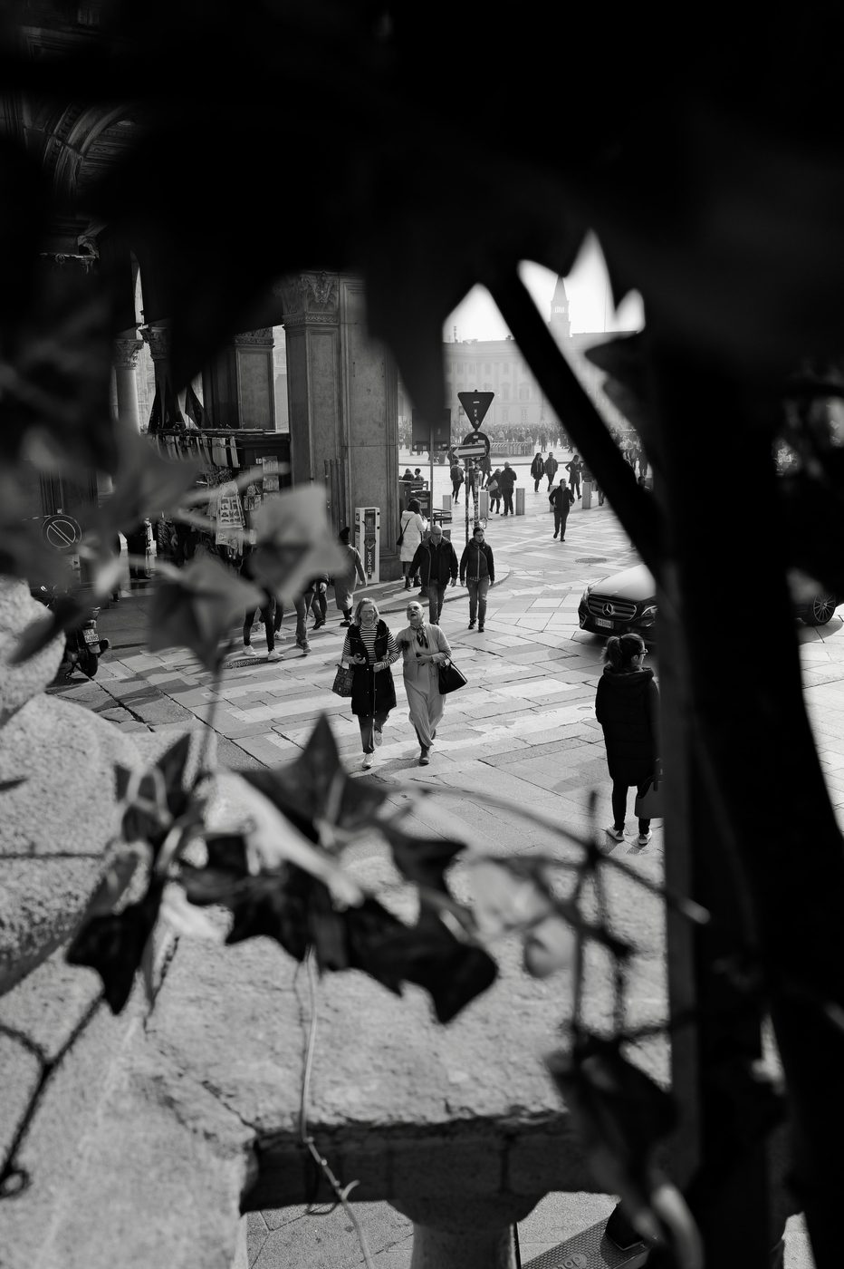 View through foliage of costumed passers-by strolling across the square, Milan