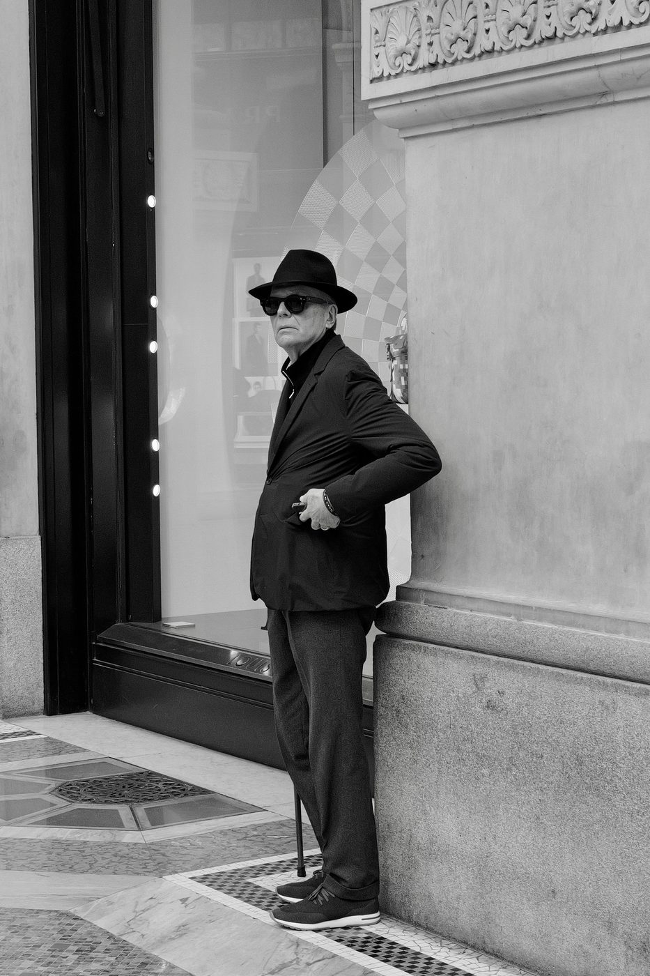 Elegant man in a hat and dark sunglasses leaning on his cane near a shop window, Milan