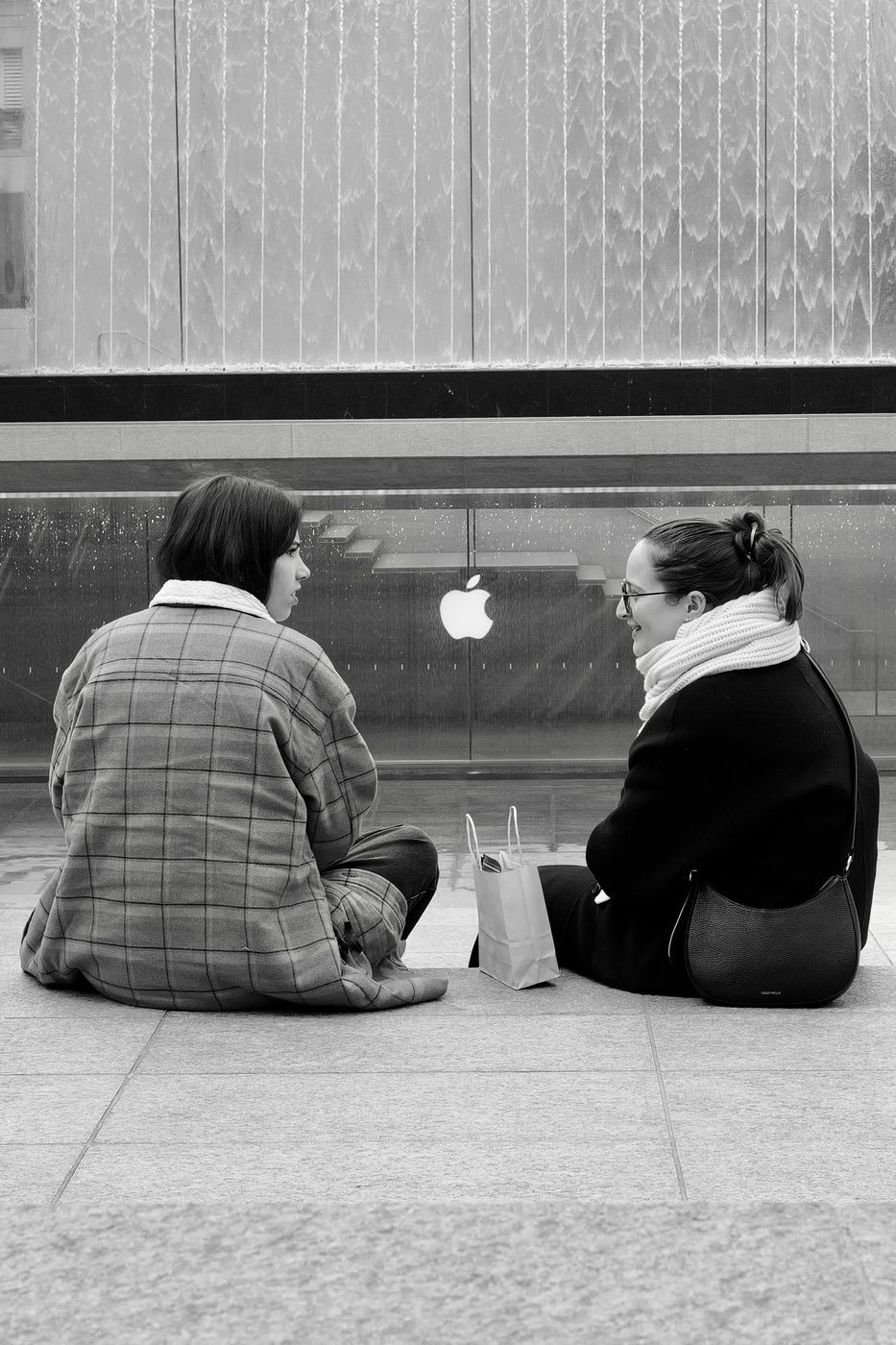 Two friends sitting with their backs to the camera in front of the Apple Store's water wall, Milan