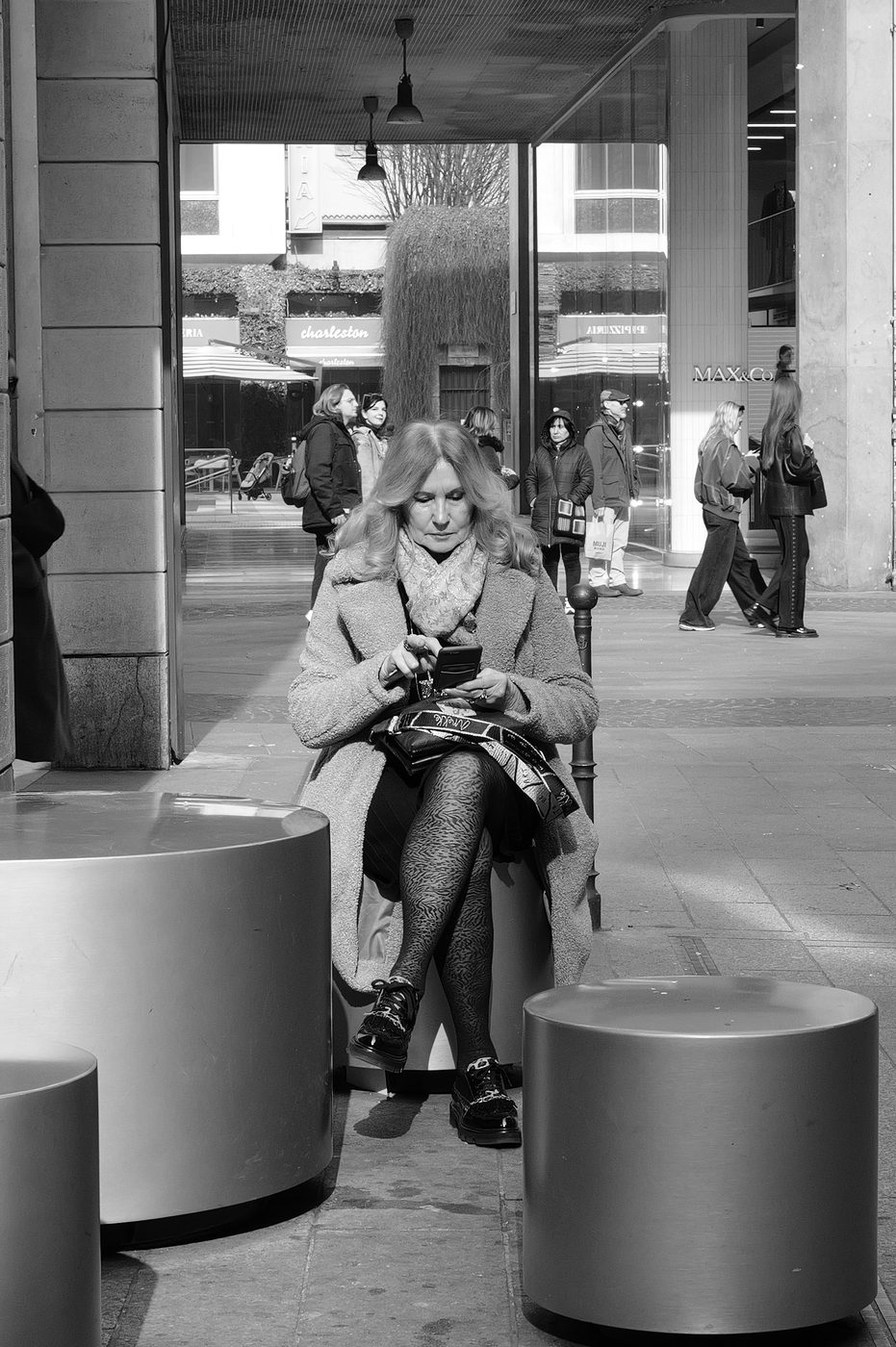 Woman sitting on a cylindrical stool checking her phone on a shopping street, Milan