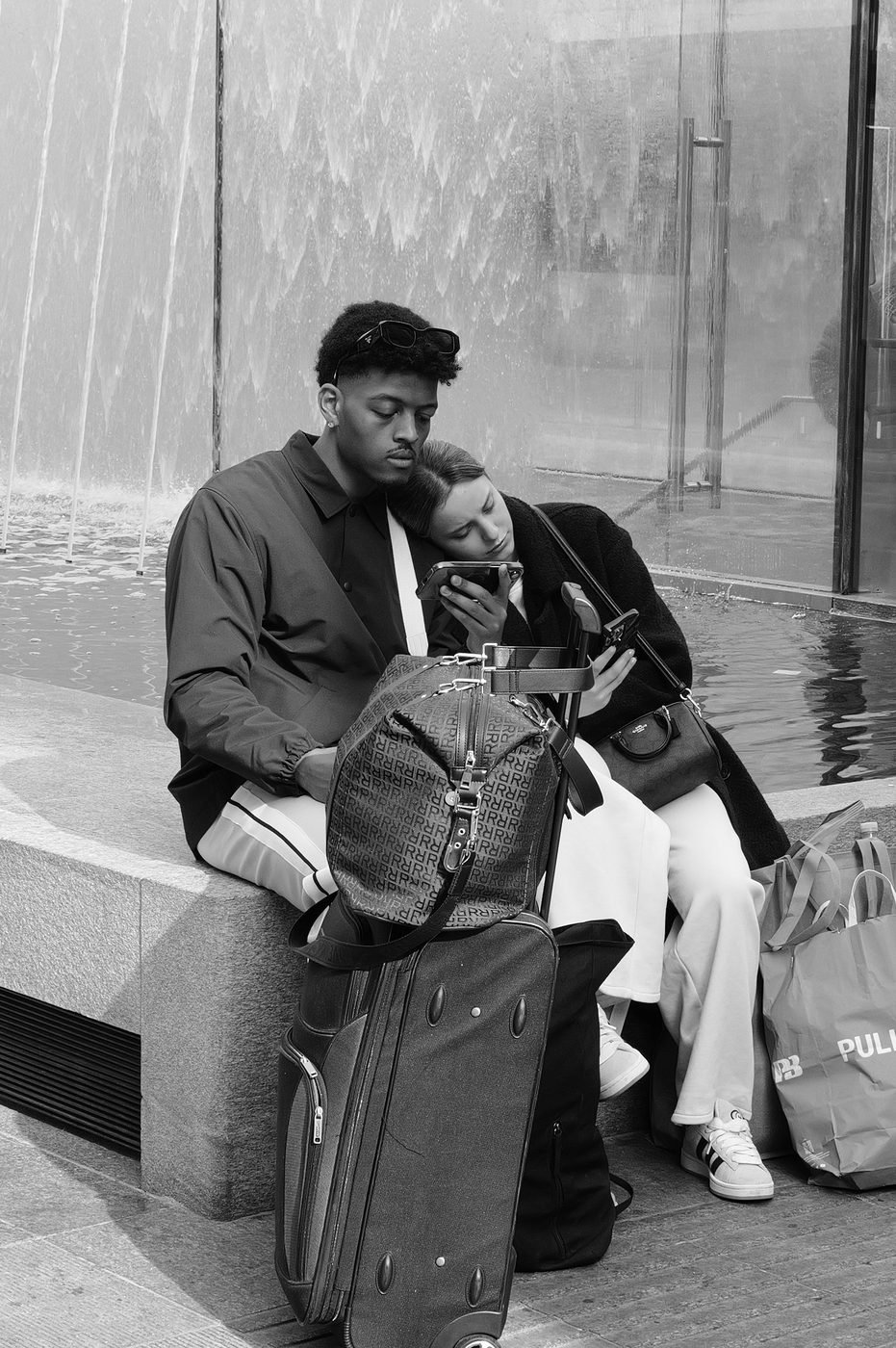 Young couple looking at a phone, suitcases set down in front of a water wall, Milan