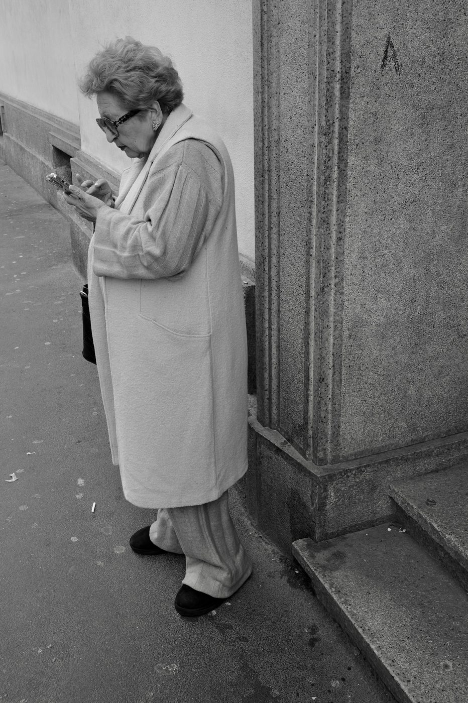 Elderly woman in a light-coloured coat checking her phone, leaning against a stone wall, Milan