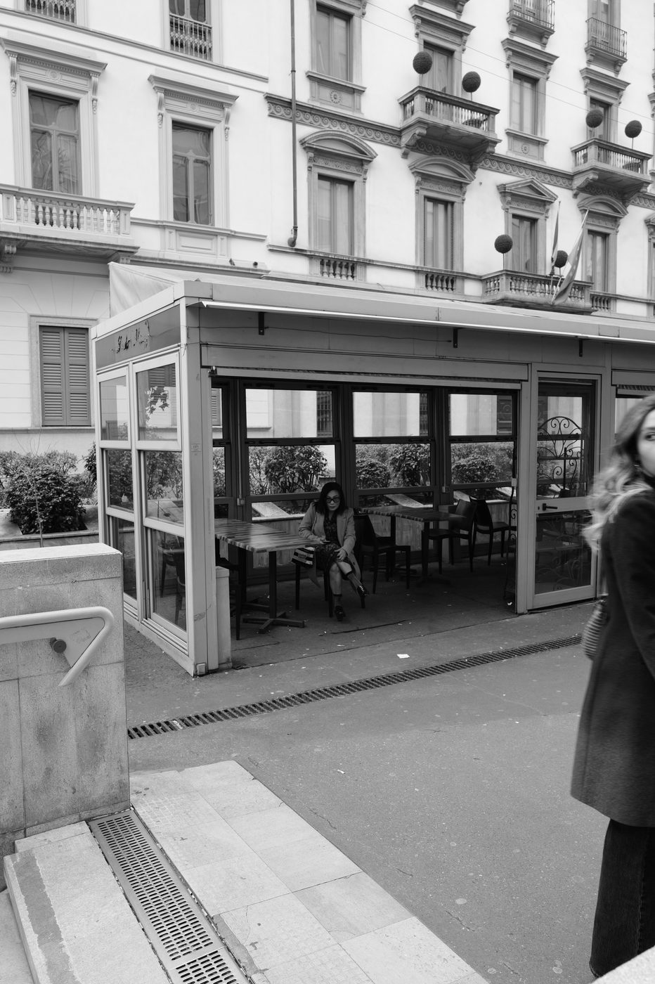 Woman sitting alone in a restaurant's glass veranda, a passer-by turning around outside, Milan